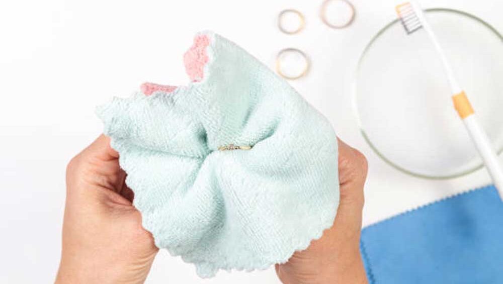 Hands gently polishing a gold-filled ring with a soft cleaning cloth, with additional cleaning tools and rings in the background.