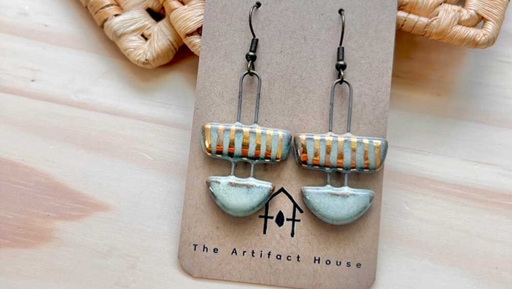 Handcrafted ceramic earrings with geometric shapes, featuring earthy tones and subtle metallic accents, displayed on a branded card from The Artifact House.