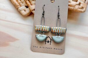 Handcrafted ceramic earrings with geometric shapes, featuring earthy tones and subtle metallic accents, displayed on a branded card from The Artifact House.