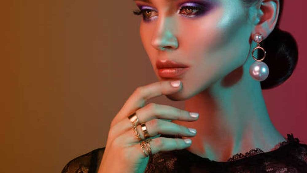 Elegant woman showcasing dynamic jewelry with bold pearl drop earrings and layered gold rings against a vibrant gradient background.