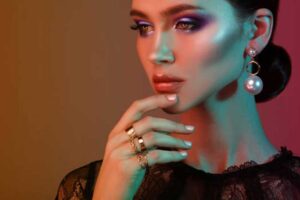 Elegant woman showcasing dynamic jewelry with bold pearl drop earrings and layered gold rings against a vibrant gradient background.