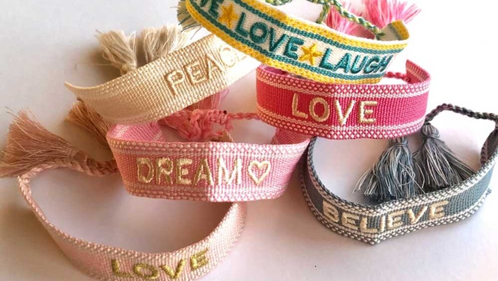 Colorful woven fabric bracelets with tassels, featuring words like 'Peace,' 'Love,' 'Dream,' 'Believe,' and 'Live Laugh Love' embroidered on them.
