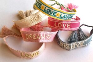 Colorful woven fabric bracelets with tassels, featuring words like 'Peace,' 'Love,' 'Dream,' 'Believe,' and 'Live Laugh Love' embroidered on them.
