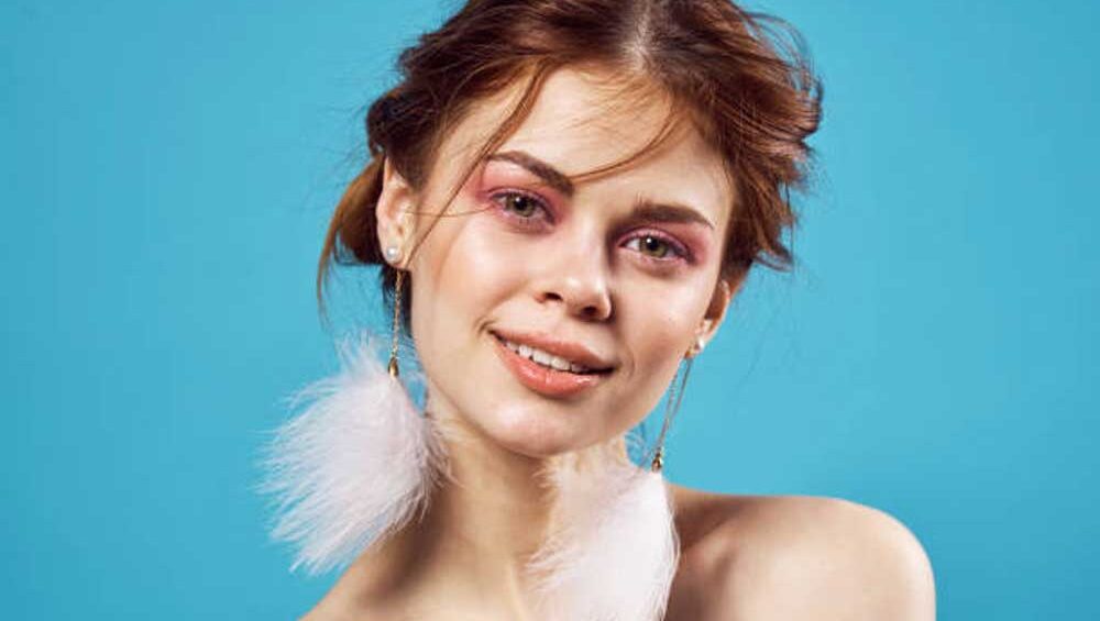 A young woman with a cheerful expression wearing statement white feather earrings against a vibrant blue background.