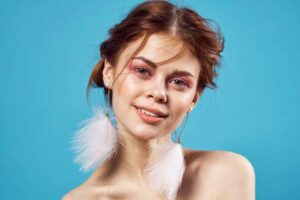 A young woman with a cheerful expression wearing statement white feather earrings against a vibrant blue background.