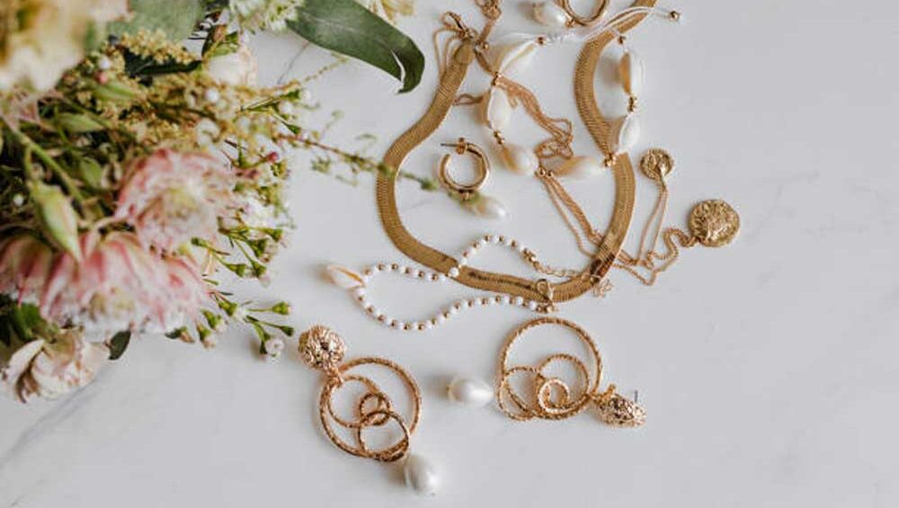 A stylish arrangement of mix-and-match jewelry pieces, featuring gold necklaces, rings, and earrings alongside delicate flowers on a white marble surface.