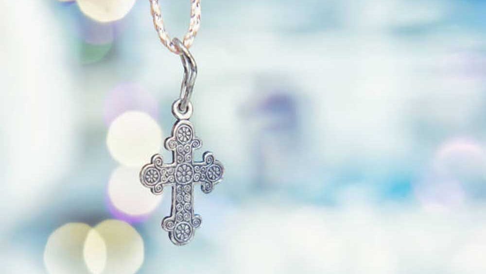 A silver cross pendant with intricate details hanging on a chain, against a soft, blurred background of light bokeh.