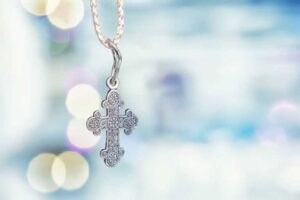 A silver cross pendant with intricate details hanging on a chain, against a soft, blurred background of light bokeh.