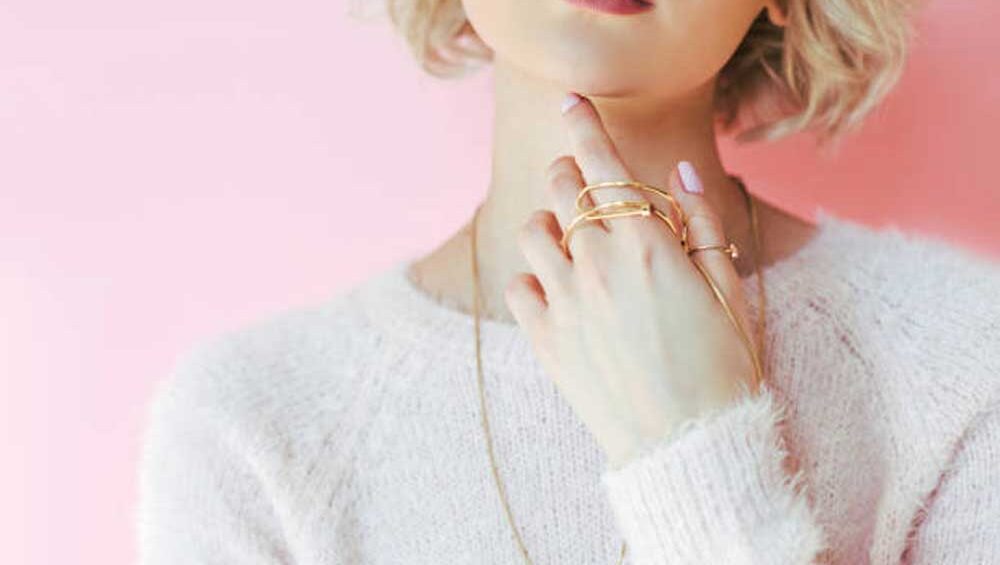 Close-up of a woman with short blonde hair wearing gold rings, necklaces, and a fuzzy pink sweater against a soft pink background.