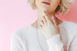Close-up of a woman with short blonde hair wearing gold rings, necklaces, and a fuzzy pink sweater against a soft pink background.