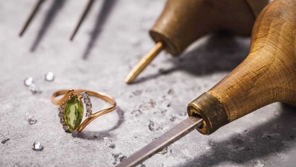 A handcrafted gold ring with a green gemstone and diamond accents placed on a workbench surrounded by artisan jewelry tools.