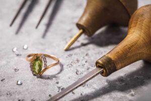 A handcrafted gold ring with a green gemstone and diamond accents placed on a workbench surrounded by artisan jewelry tools.