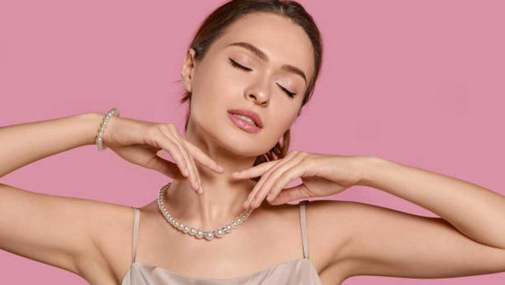 Elegant woman wearing a pearl necklace and bracelet, posing gracefully with her hands near her face against a soft pink background.