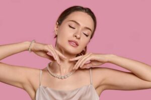 Elegant woman wearing a pearl necklace and bracelet, posing gracefully with her hands near her face against a soft pink background.