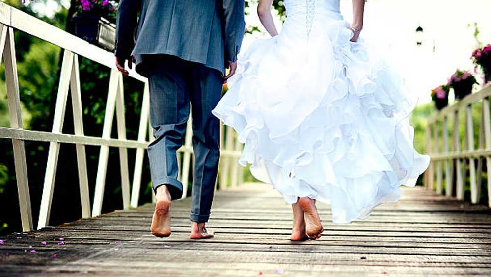 A newlywed couple walks barefoot on a rustic wooden bridge, symbolizing grounding, connection, and a love rooted in nature.