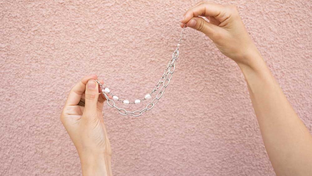 Hands gently holding a silver bracelet with a layered chain design against a textured pink wall background.