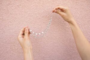 Hands gently holding a silver bracelet with a layered chain design against a textured pink wall background.