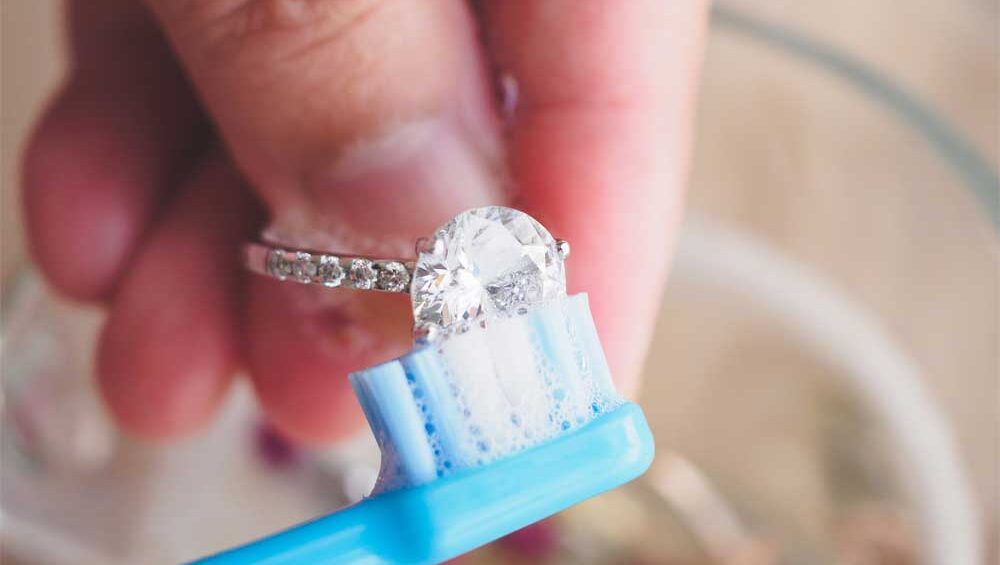 Close-up of a diamond ring being gently cleaned with a toothbrush, showing the process of jewelry care.