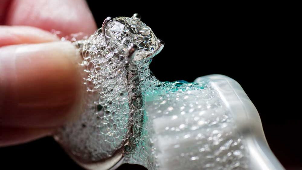 Close-up of a silver ring being cleaned with a toothbrush and soapy bubbles.