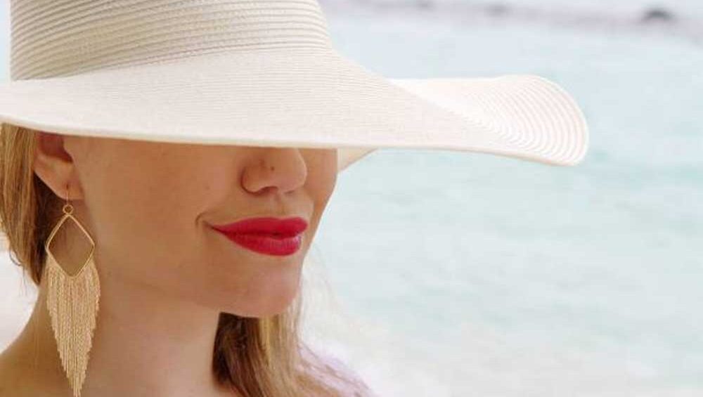 Close-up of a woman wearing a wide-brimmed white hat, red lipstick, and gold fringe earrings at the beach.