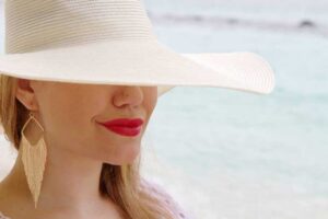 Close-up of a woman wearing a wide-brimmed white hat, red lipstick, and gold fringe earrings at the beach.