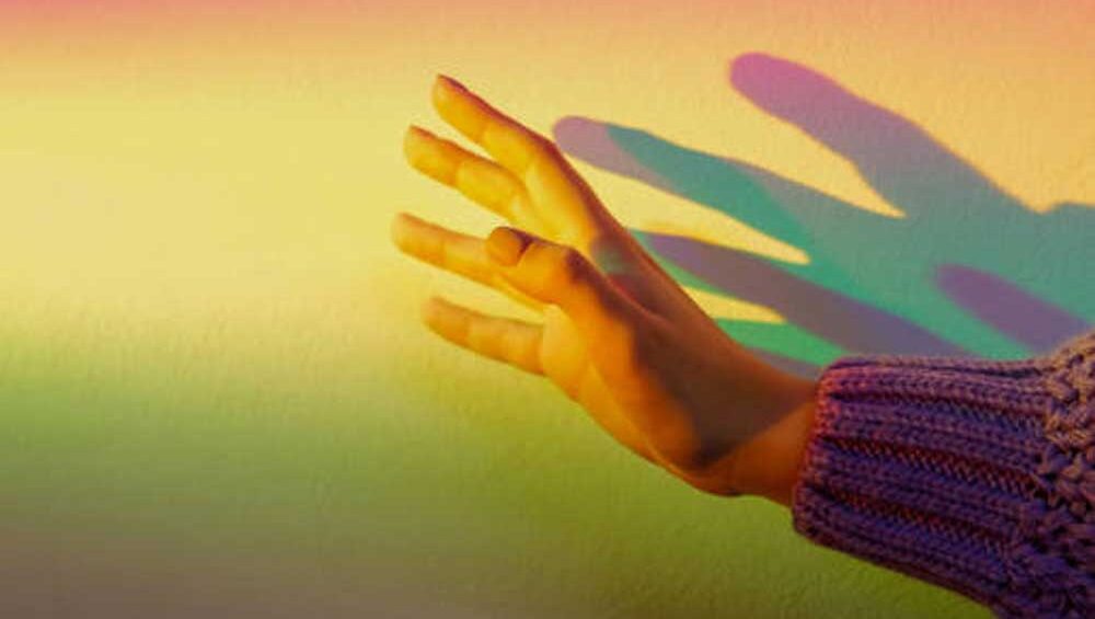 A softly lit hand reaching toward a colorful gradient light on a wall, symbolizing emotional transition and the journey through love.