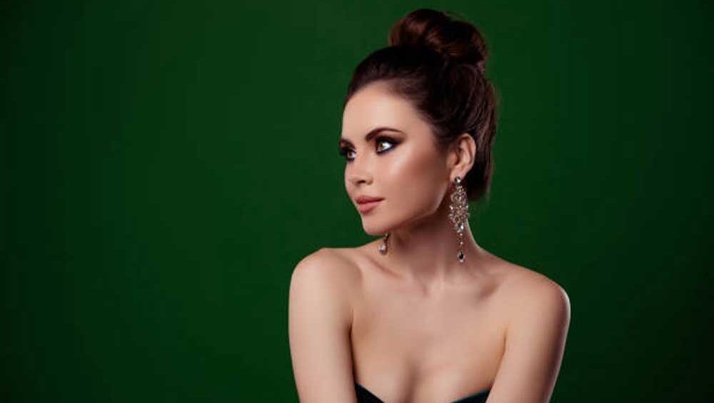 A woman in a sleek black dress with a glamorous updo, wearing bold chandelier earrings against a rich green background, exuding nighttime elegance.