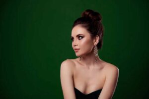 A woman in a sleek black dress with a glamorous updo, wearing bold chandelier earrings against a rich green background, exuding nighttime elegance.