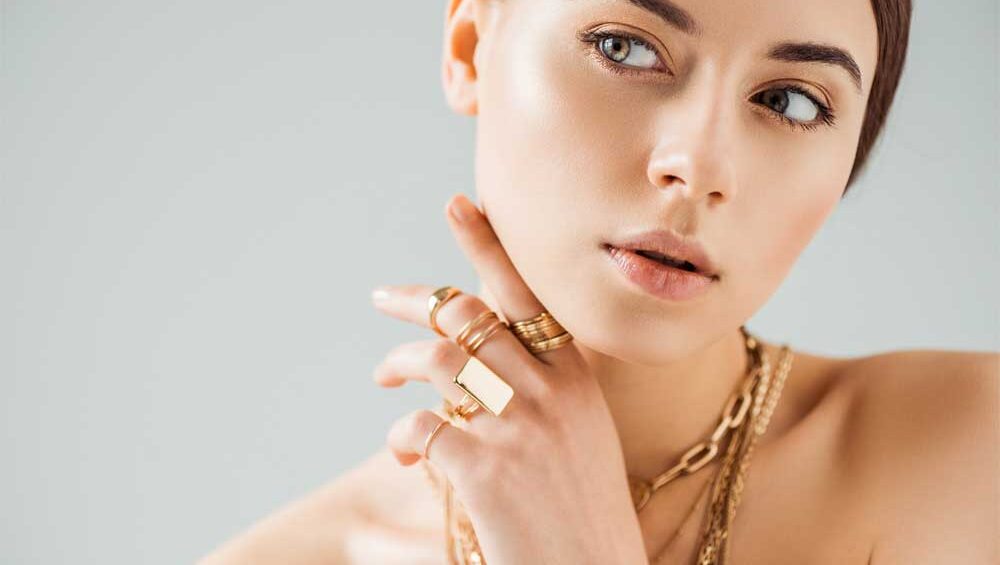 Elegant woman wearing gold jewelry, including rings and a chain necklace, posing against a neutral background with a polished and sophisticated look.