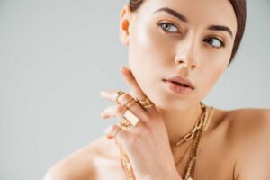 Elegant woman wearing gold jewelry, including rings and a chain necklace, posing against a neutral background with a polished and sophisticated look.