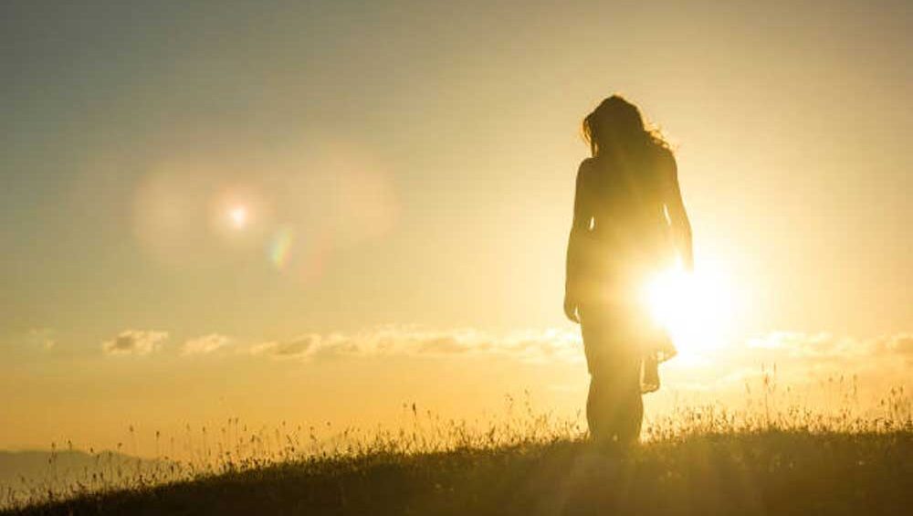 Silhouette of a young woman in a dress standing on grass against a mountain sunset, symbolizing empowerment and transformation.
