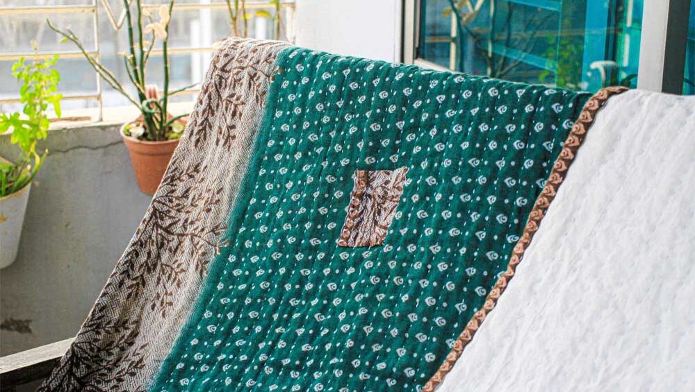 Hand-stitched Kantha quilt with green, white, and patterned fabric draped over a chair on a sunlit balcony.