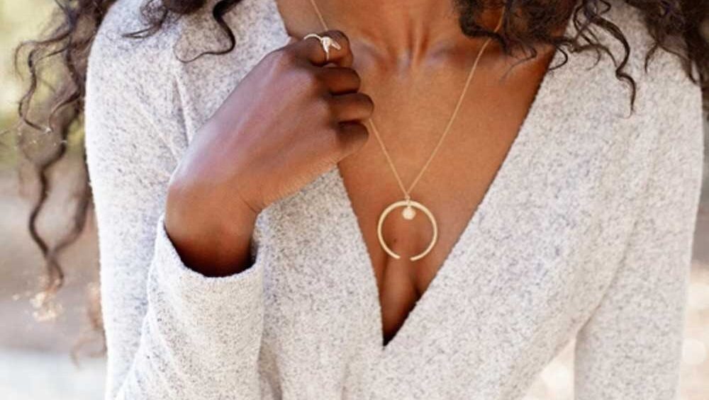 Close-up of a woman wearing a minimalist gold circle pendant necklace and a delicate ring, styled with a light gray V-neck top.