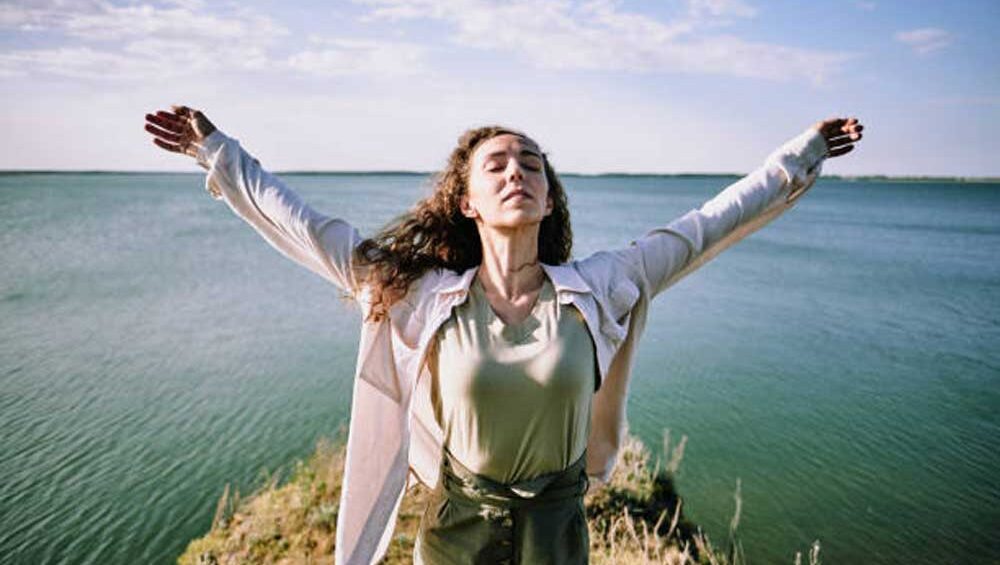 A woman stands by the water with her arms open wide, eyes closed, embracing freedom and inner peace.