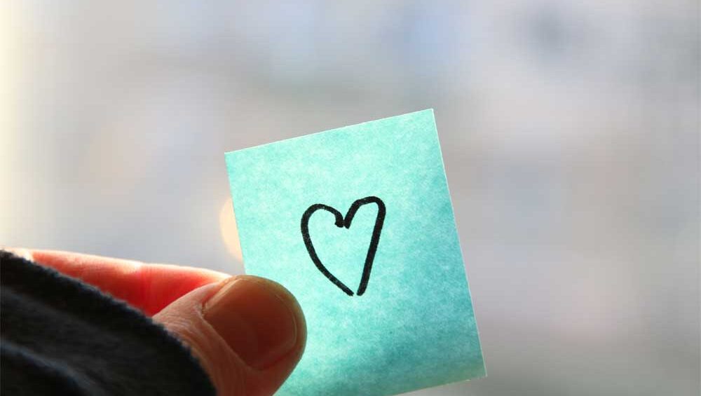 A hand holding a small blue sticky note with a simple hand-drawn heart, symbolizing love and self-worth.