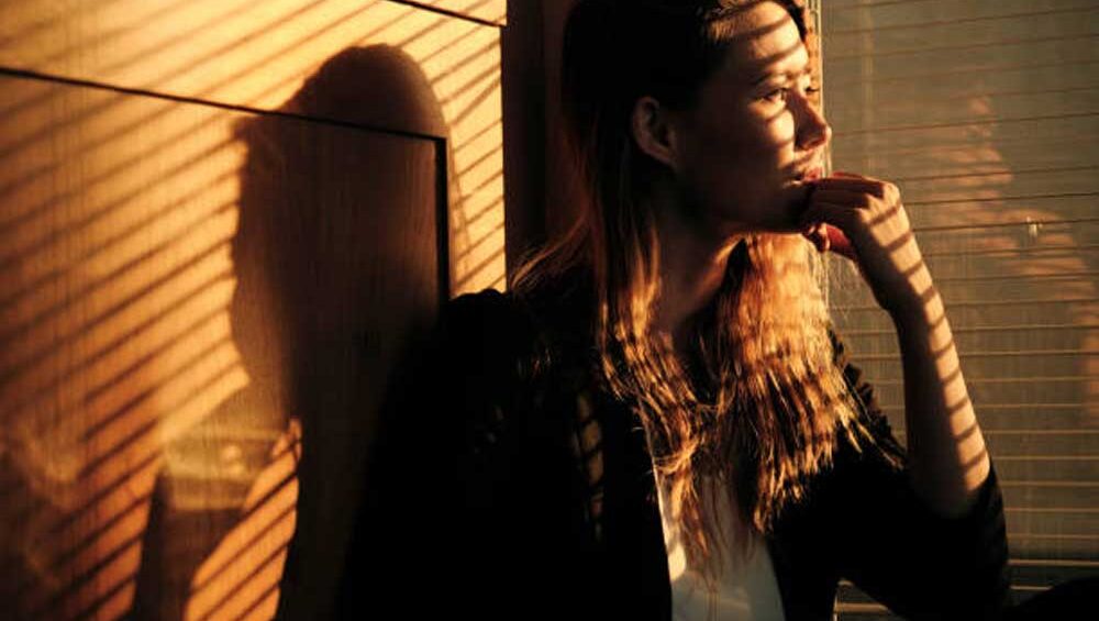 A woman sits by a window, deep in thought as golden light and shadows create an introspective atmosphere, symbolizing emotional depth and self-discovery.