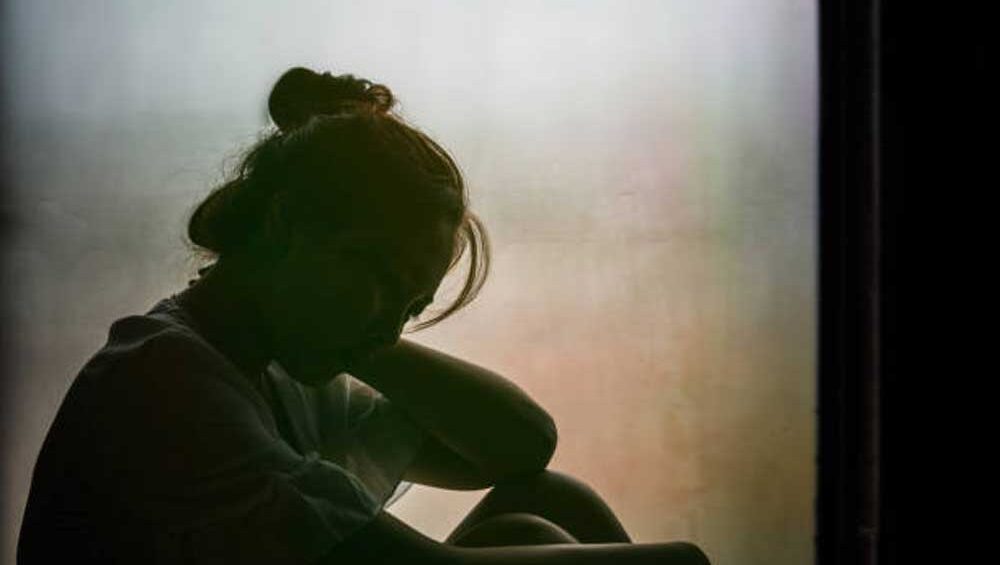 A woman sitting by a window, deep in thought, looking troubled and distant, representing the emotional weight of distrust and misunderstanding in family relationships.