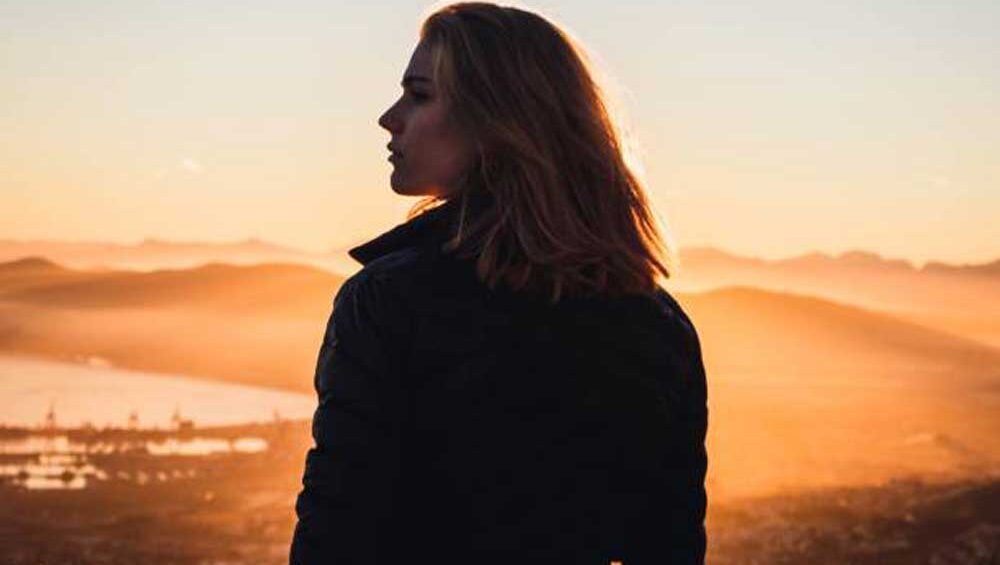 A woman stands at the edge of a hill, gazing into the horizon as the sun sets, casting a golden glow. Her silhouette reflects quiet strength and introspection.
