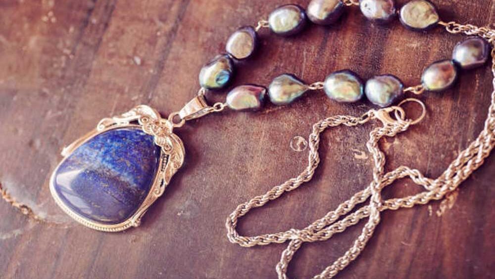 Close-up of a lapis lazuli pendant necklace with baroque pearls and a gold chain on a rustic wooden surface.