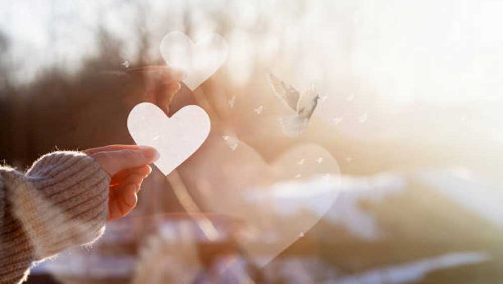 A hand holding a paper heart with soft sunlight filtering through, symbolizing love, hope, and new beginnings. A dove and glowing hearts add to the dreamy, ethereal atmosphere.