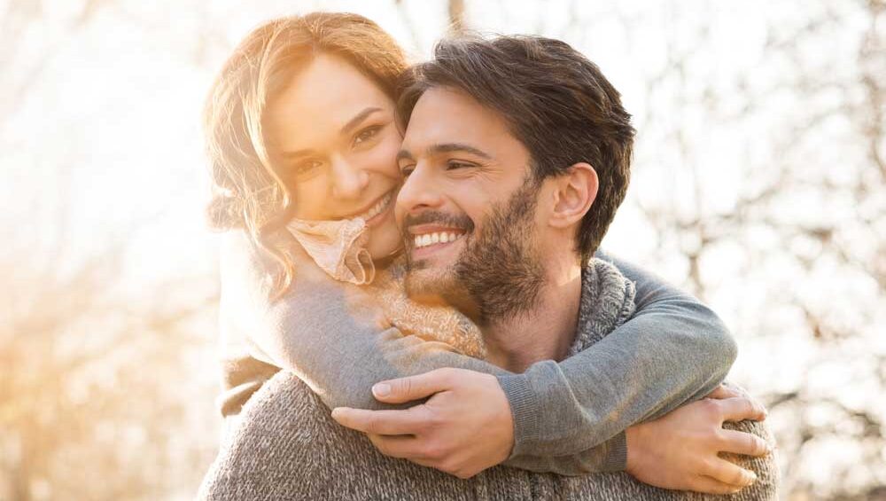 Woman embracing man from behind while both smile warmly, bathed in golden sunlight