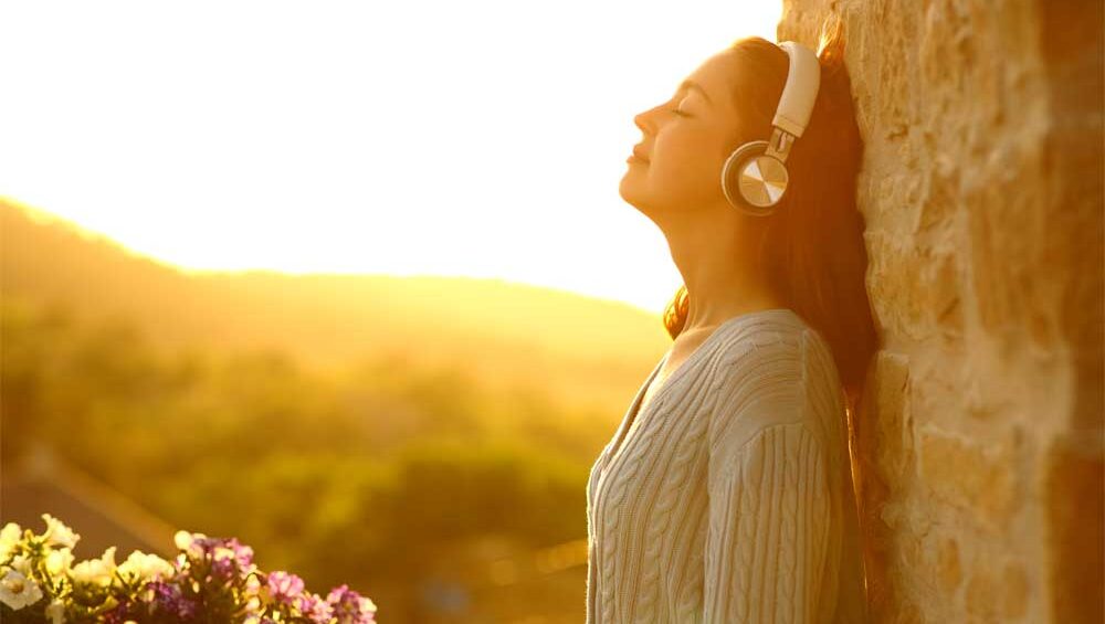 Woman wearing headphones, eyes closed, leaning against a sunlit stone wall while listening to music, symbolizing peace, reflection, and self-discovery.
