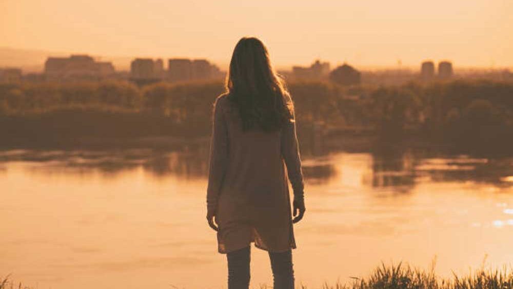 A woman stands alone by the water at sunset, gazing into the distance, reflecting on her emotions.