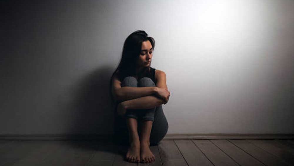 A woman sitting alone against a wall, hugging her knees, looking down in deep contemplation, conveying sadness and emotional distance.