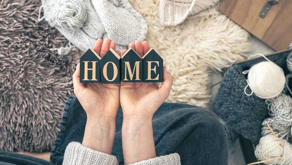 Hands holding small wooden blocks spelling "HOME" surrounded by cozy textures like yarn, knits, and rugs.