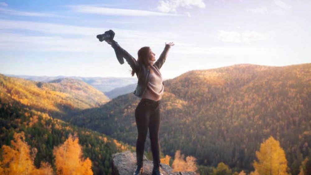 A woman stands on the edge of a cliff with arms raised in triumph, symbolizing freedom, strength, and breaking free from limitations.