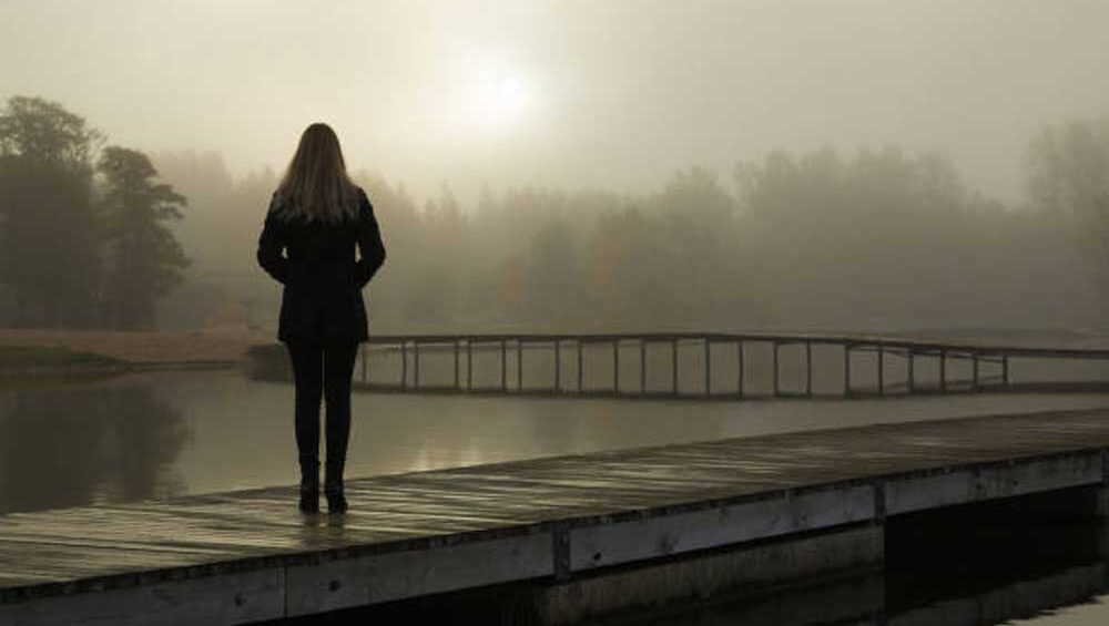 A solitary woman stands on a wooden dock overlooking a misty lake, surrounded by fog, evoking a sense of introspection and deep emotion.