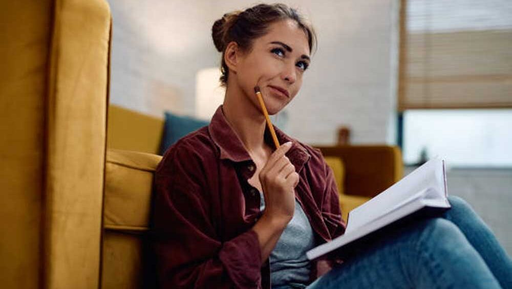 A thoughtful young woman sitting on the floor with a notebook, reflecting deeply while holding a pencil.