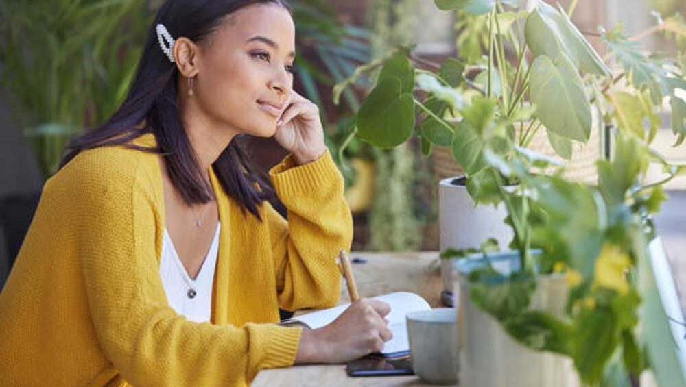 Young woman journaling near plants, reflecting with a peaceful expression, symbolizing emotional intelligence and self-awareness.