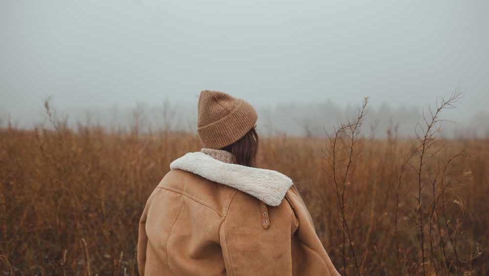 Woman in a brown coat standing in a misty field, symbolizing the emotional weight we quietly carry.